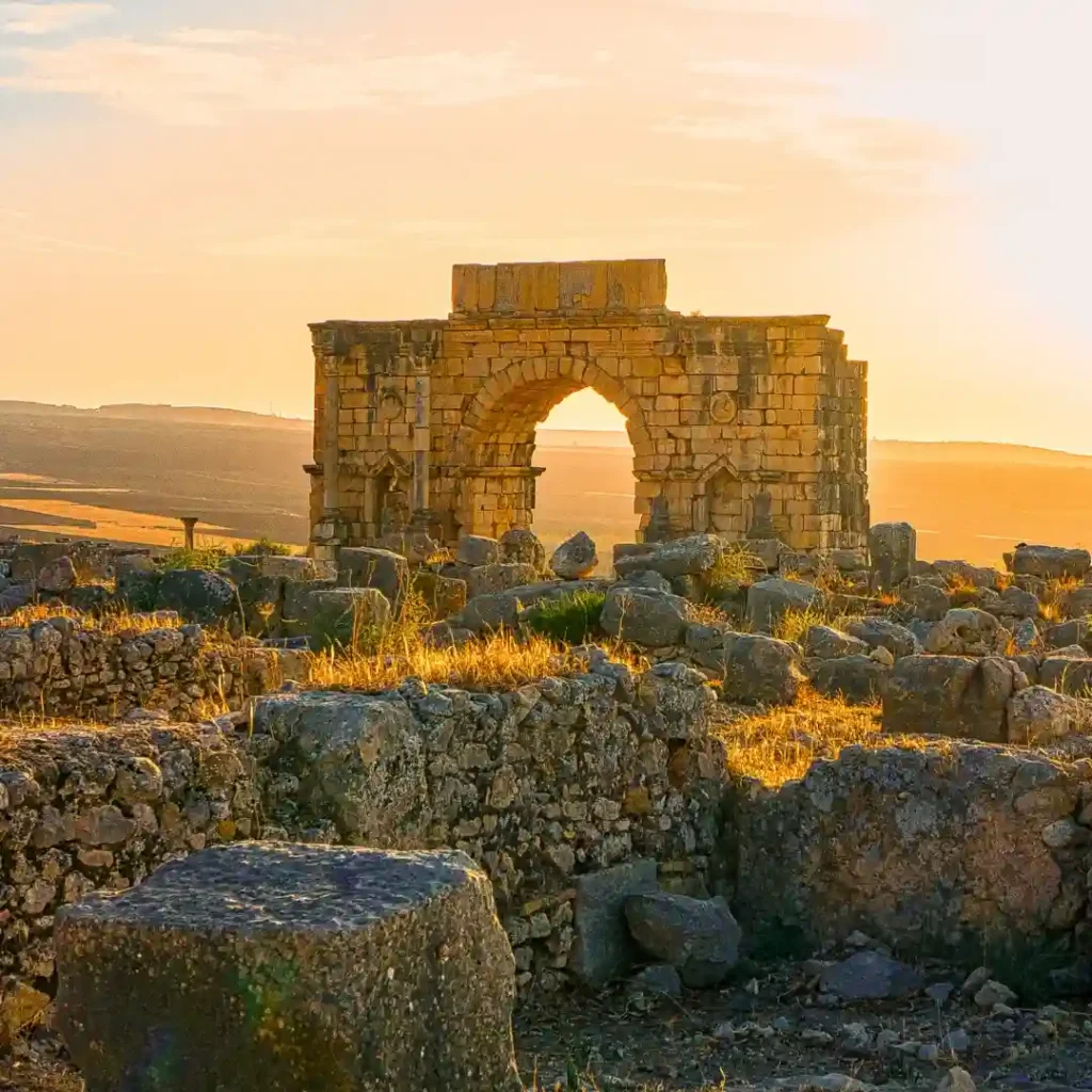 UNESCO-listed Roman ruins of Volubilis with stone columns, arches, and ancient mosaics under the Moroccan sun. Visit Morocco Tours.