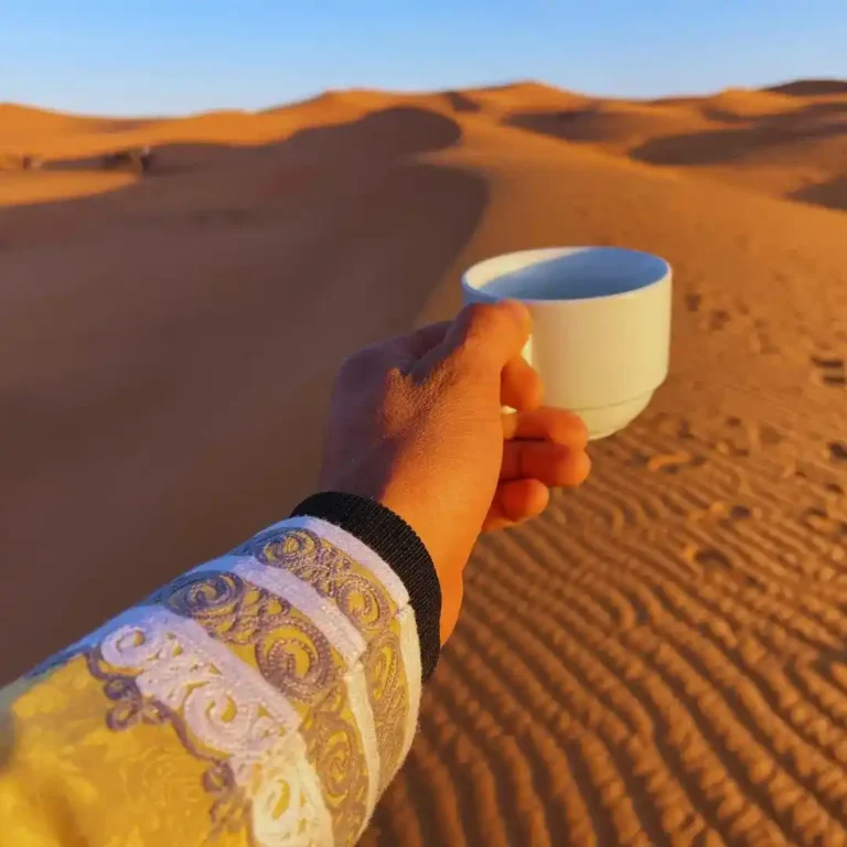 A hand holding a traditional Moroccan coffee with a view of the golden Erg Chebbi sand dunes at sunset. Visit Morocco Tours